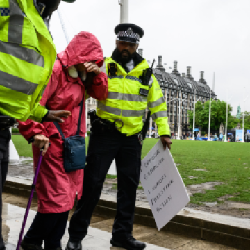 a person using a cane is led away by two police men in front of the big ben for holding a placard. text reads "end prosecution of peaceful protestors" a person using a cane is led away by two police men in front of the big ben for holding a placard. text reads "end prosecution of peaceful protestors"