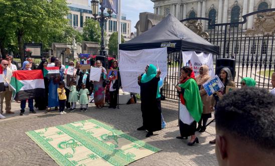Vigil for peace, justice and accountability for war crimes in Sudan - Belfast, June 2023. Image shows speaker and supporters, many holding Sudan flags, in front of Belfast City Hall