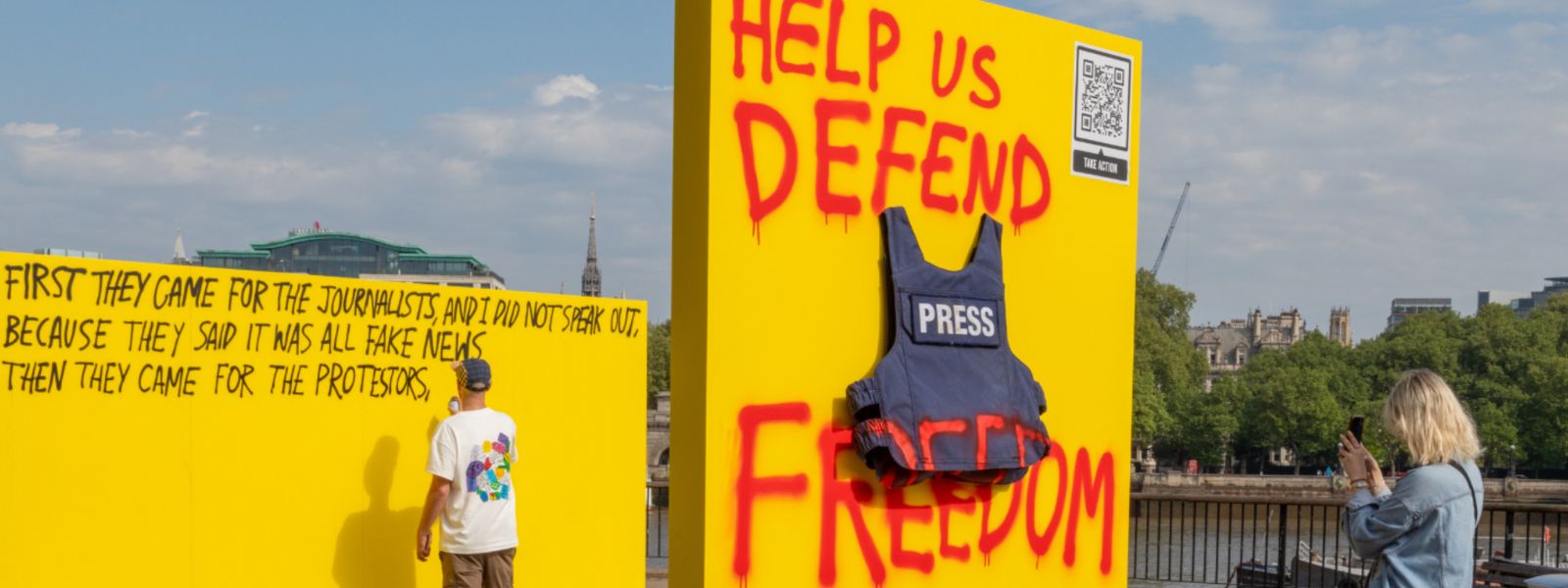 Stunt in London by the river with an artist writing a poen about protest rights, a yellow wall with a press jacket and graffiti saying help us defend freedom, and a woman taking photo of the scene Stunt in London by the river with an artist writing a poen about protest rights, a yellow wall with a press jacket and graffiti saying help us defend freedom, and a woman taking photo of the scene