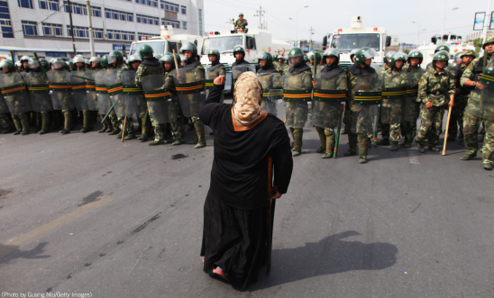 "A person stands in front of a line of heavily armed soldiers who have their shields up. The person has their first in the air in defiance'