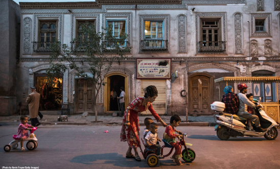 "A street in North East China at nighttime shows a scene of a woman helping three children play on a small bike. Another chlld on a toy bike is behind them. The buildings are partially lit by what looks like a street light out of shot. There is also a person on a moped passing by the children""