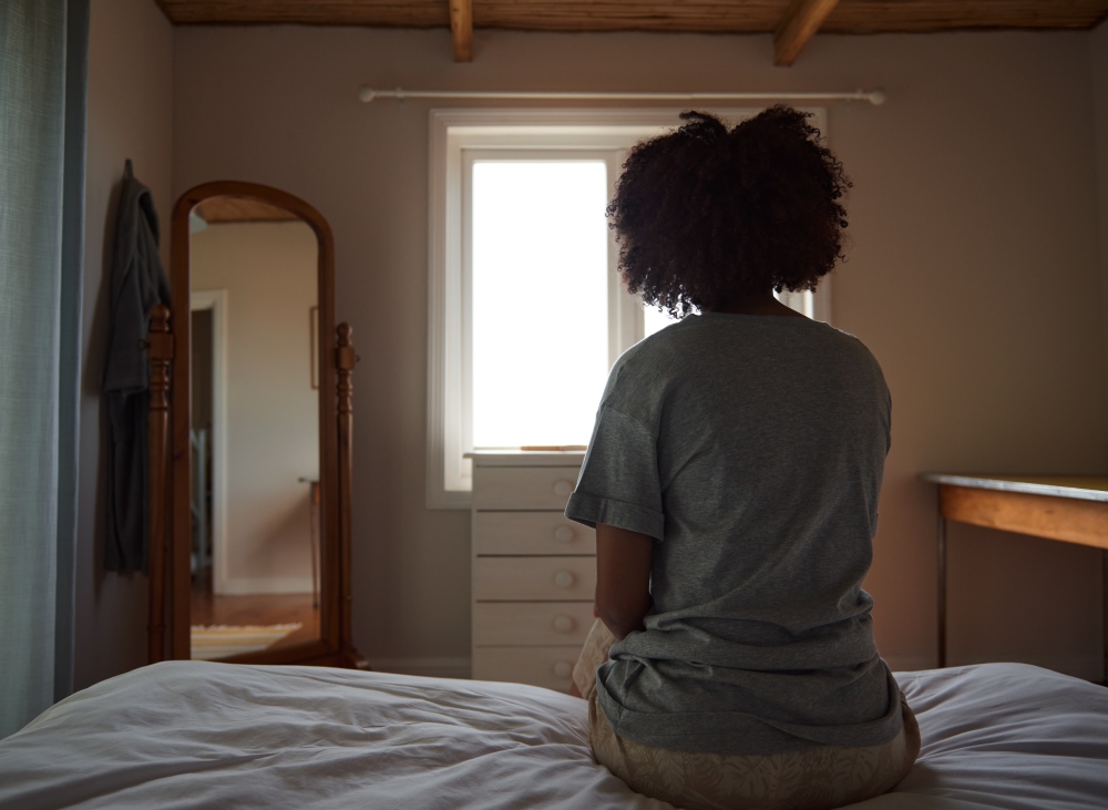 Rear view of a woman suffering from depression sitting on her bed and looking out through a window
