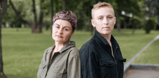 Sphere NGO founders, Anna and Vira stand back to back outside in a park and look at camera