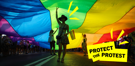 A person stands under a rainbow flag during a protest