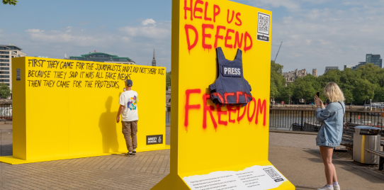 Photo of a press jacket pinned to a yellow board. Red spray paint reads 'Help us defend press freedom'