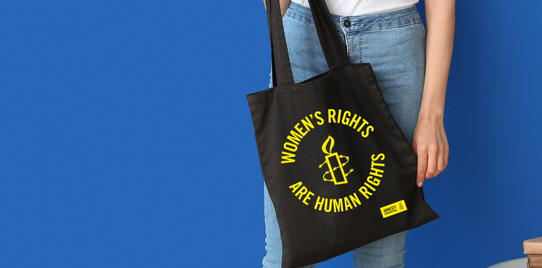 Woman holds black tote bag that has the text 'women's rights are human right's' and the Amnesty International logo in yellow