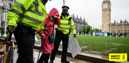 a person using a cane is led away by two police men in front of the big ben for holding a placard. text reads "end prosecution of peaceful protestors"