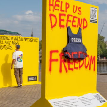 Photo of a press jacket pinned to a yellow board. Red spray paint reads 'Help us defend press freedom'