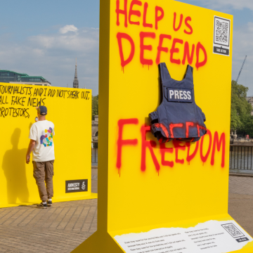 Photo of a press jacket pinned to a yellow board. Red spray paint reads 'Help us defend press freedom'