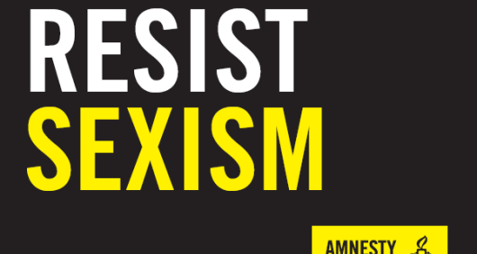 A black placard that reads 'Resist Sexism' with an Amnesty logo in the bottom-right corner