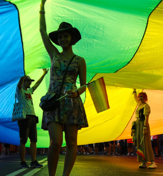 person under a pride flag tent, holding a pride flag, holding it up in protest