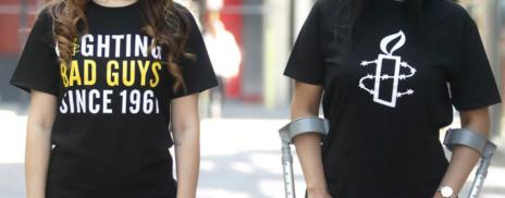 Two people wearing Amnesty T-shirts, one with the 'Fighting Bad Guys Since 1961' and the other with the classic Candle design.