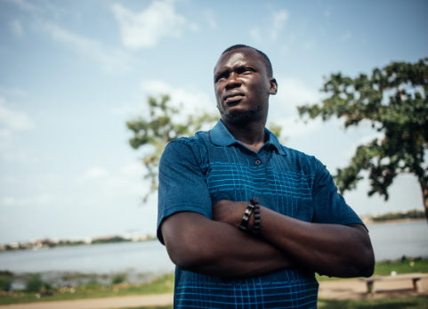 Imoleayo stands outside with his arms folded looking away from camera, wearing a blue shirt