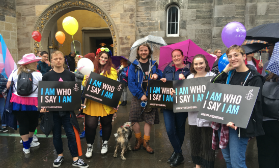 A group of six individuals from Amnesty International Scotland holding black signs with the text 'I Am Who I Say I Am' printed in pink and blue.