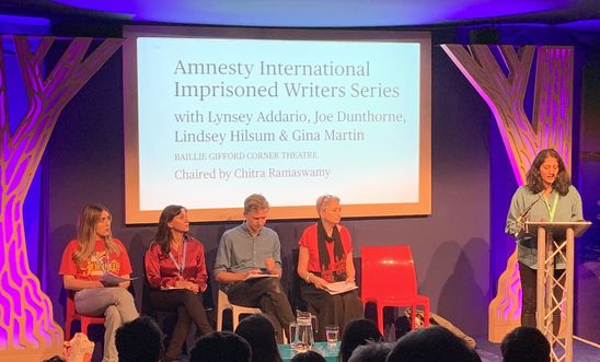 Image shows a woman reading at a lectern whilst four people sit on a stage 