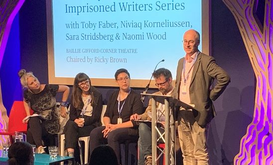 Image shows a man reading at a lectern whilst four people sit on a stage 