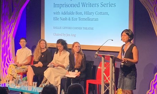 Image shows a woman reading at a lectern whilst four people sit on a stage 