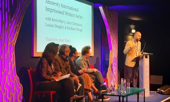 Image shows a man reading at a lectern whilst four people sit on a stage Image shows a man reading at a lectern whilst four people sit on a stage