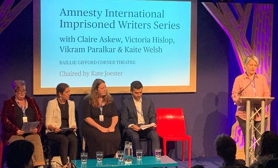 Image shows a woman reading at a lectern whilst four people sit on a stage 