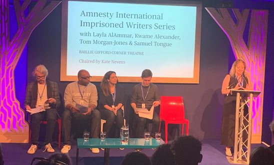 Image shows a woman reading at a lectern whilst four people sit on a stage 