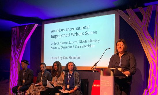Image shows a woman reading at a lectern whilst four people sit on a stage Image shows a woman reading at a lectern whilst four people sit on a stage