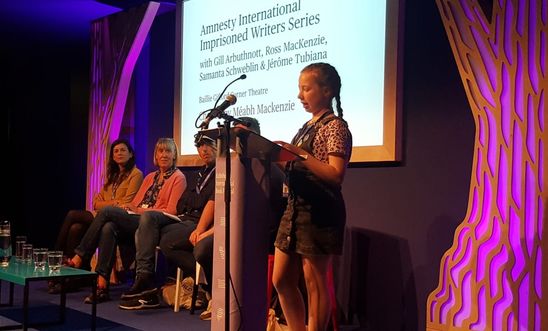 Image shows a girl reading at a lectern whilst four people sit on a stage 