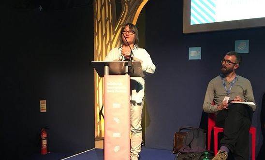 Image shows a woman reading at a lectern whilst a man looks on admiringly Image shows a woman reading at a lectern whilst a man looks on admiringly