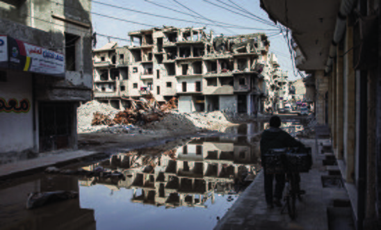 Raqqa, Syria - A silhouetted man with his bike facing the ruins of a large building which is also reflected in a pool of water