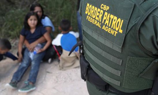 Woman sitting on ground with two children with border patrol agent in foreground