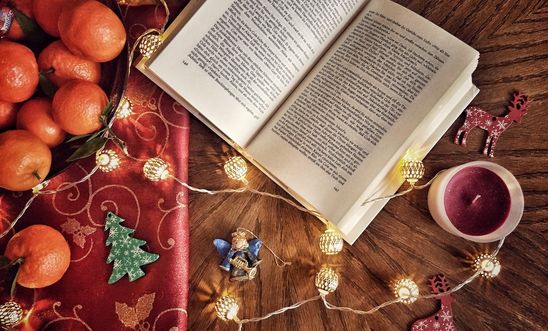 An open novel nestled next to a class of mulled wine, on a table with satsumas and gift wrapping trinkets.