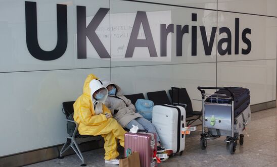Two people in full PPE in a waiting area with their luggage with a 'UK Arrivals' sign behind them