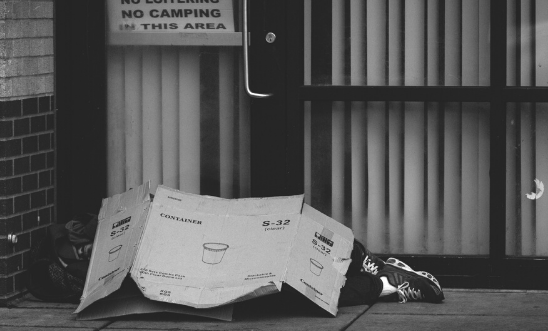 A person lies beneath cardboard on the ground outside, image inbalck and white