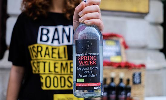 Image includes person with a shirt that reads 'ban israeli settlement goods' holding a bottle that reads: Israeli settlement spring water, too good for locals so we stole it"