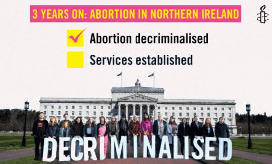 Image shows activists in front of Stormont holding signs that say DECRIMINALISE. At the top right, the amnesty candle. In highlighted pink, yellow text reads "3 years on: abortion in northern ireland", and underneath a ticked box with "Abortion decriminalised", and under it, an unticked circled box "Services established"