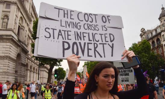 A person participating in a protest in the UK holds a placard that reads: The cost of living crisis is state inflicted poverty"