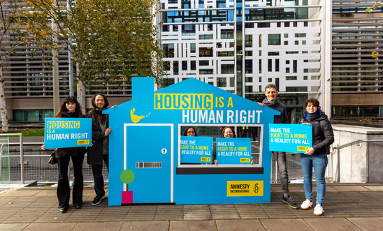 Number of people standing next to a cardboard cutout of a house saying Housing Is a Human Right Number of people standing next to a cardboard cutout of a house saying Housing Is a Human Right