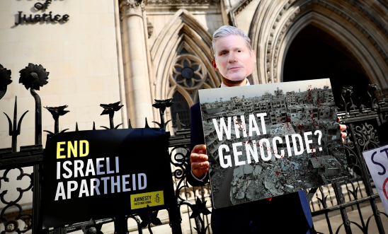 Photo featuring a masked figure of Keir Starmer holding a placard denying the mass atrocities in Gaza Photo featuring a masked figure of Keir Starmer holding a placard denying the mass atrocities in Gaza