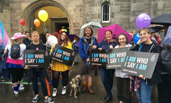 A group of six individuals from Amnesty International Scotland holding black signs with the text 'I Am Who I Say I Am' printed in pink and blue. A group of six individuals from Amnesty International Scotland holding black signs with the text 'I Am Who I Say I Am' printed in pink and blue.