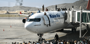 People climb on top of a standing plane at Kabul airport, and many queue beneath, hoping to board. In the background, a narrow plume of smoke billows in the distance. 