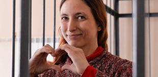 Sasha stands behind bars looking at the camera, with her hands held in a heart symbol